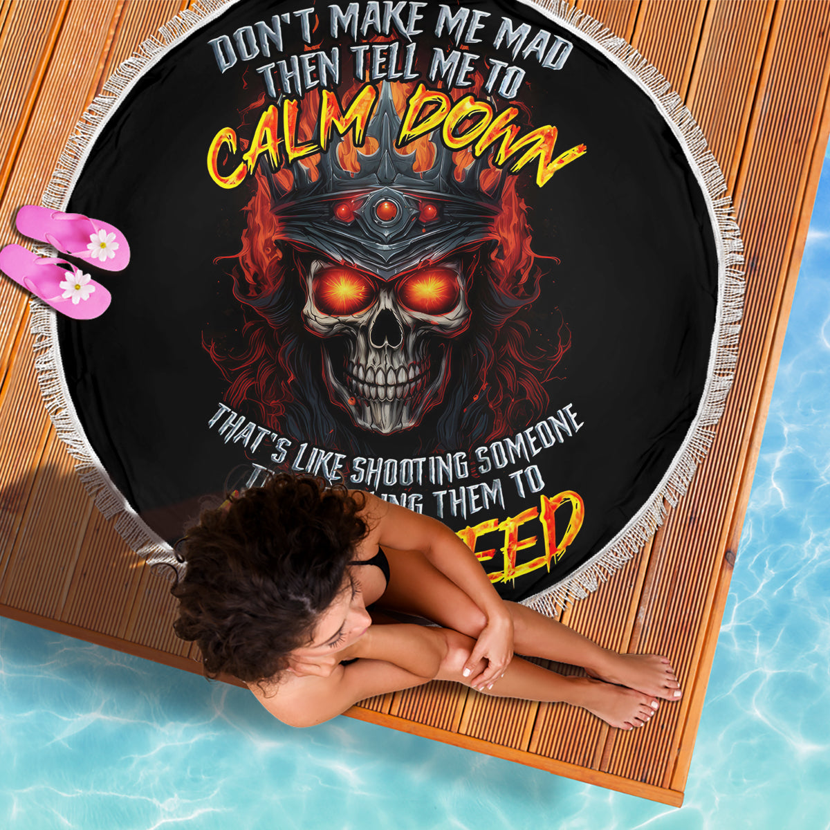 Black round beach blanket with fiery skull and bold slogan print, white fringed border; dog nearby.