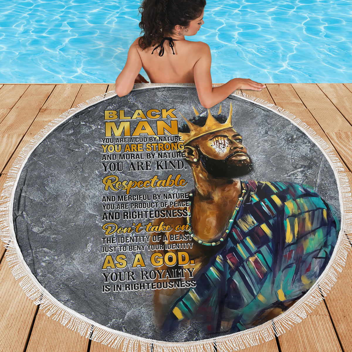 Round black beach blanket with printed portrait and gold text, fringed edge on wooden deck