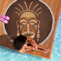Round brown beach blanket featuring a tribal mask face graphic and white fringe edge