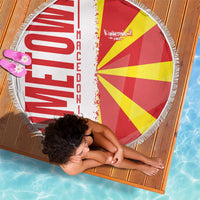Round beach blanket with red and yellow flag rays and white fringe border, laid on wood decking