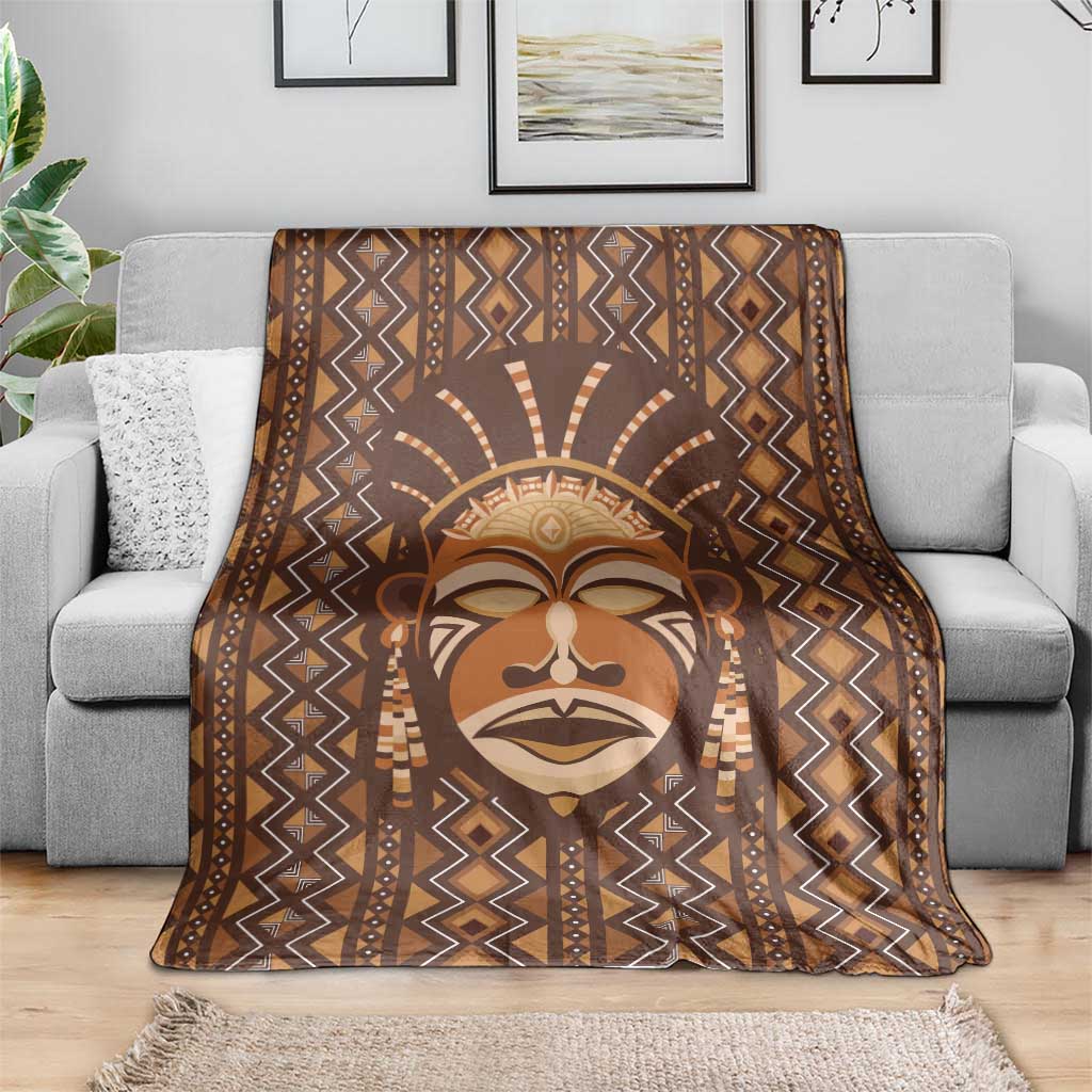 Brown mudcloth-style blanket with stylized tribal face centered and zigzag diamond pattern.