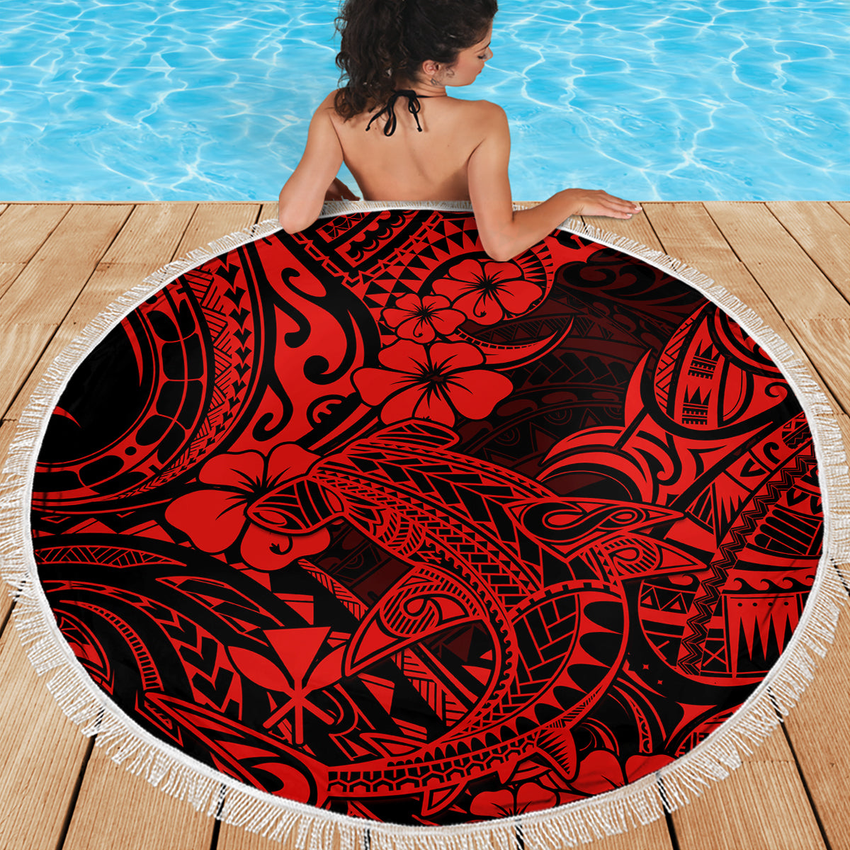 Round red beach blanket with black tribal hibiscus design and white fringe, on wooden deck