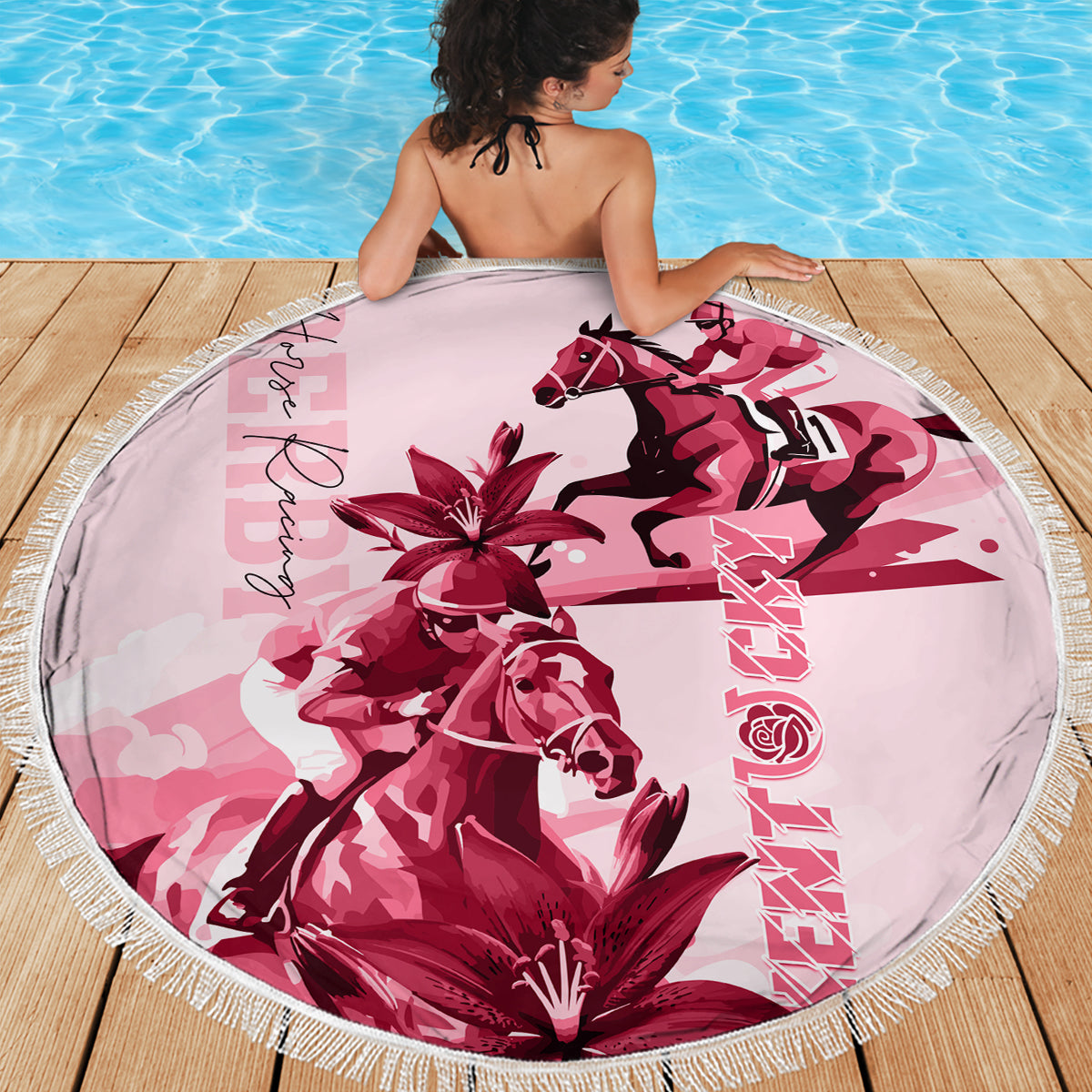 Pink round beach blanket with fringe edge, horse and lily graphic, spread on wooden deck.