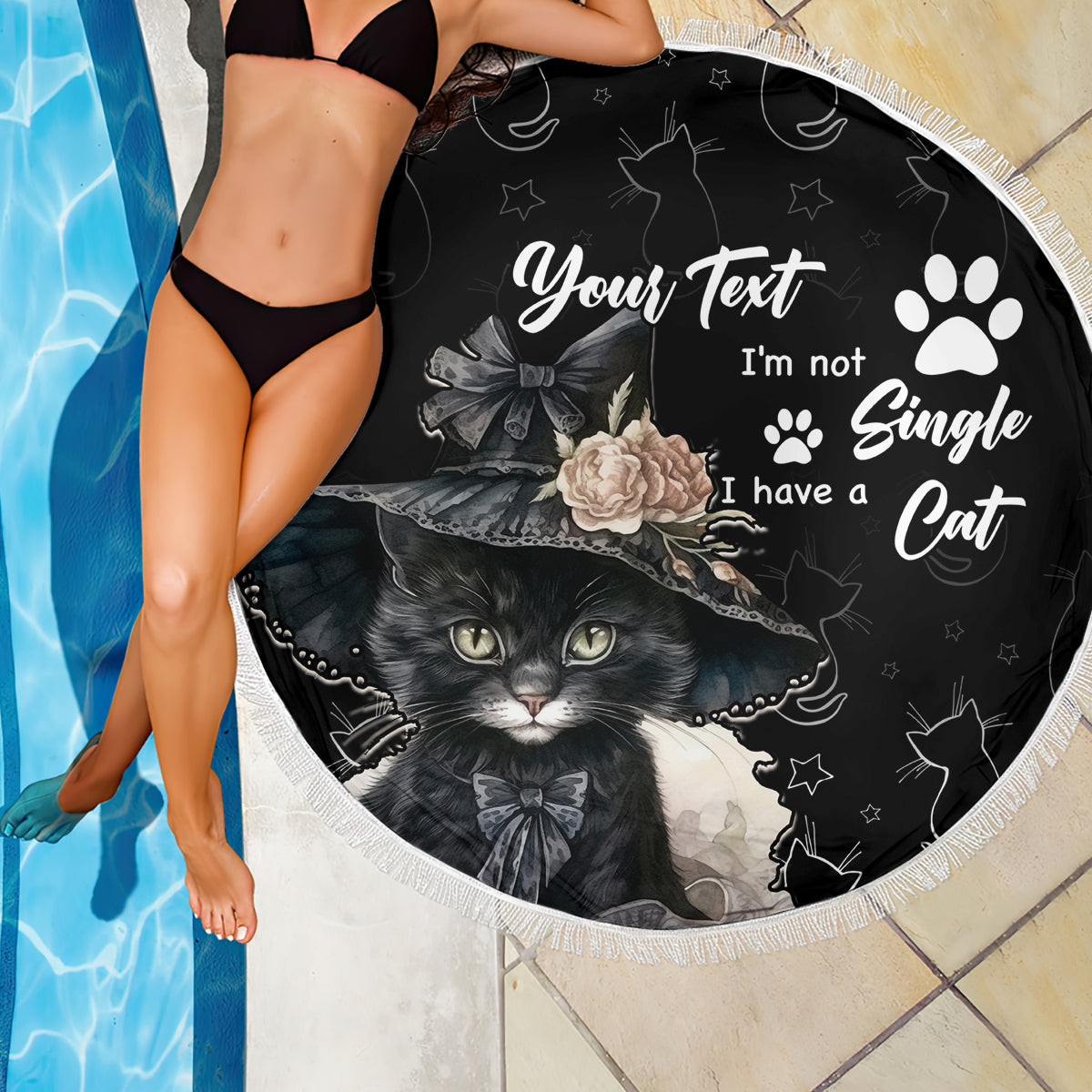 Black round beach blanket with cat graphic and white text, fringed edge over tile