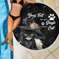 Black round beach blanket with cat graphic and white text, fringed edge over tile