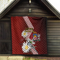 Red quilt hanging on wooden exterior with Latvian stripes, daisies, crest, and bird