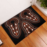 Black rubber doormat with orange and white tribal figure motifs, shown on wood floor.