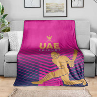 Pink UAE Cricket blanket with gold batsman illustration and dark pink chevron stripes, laid on sofa