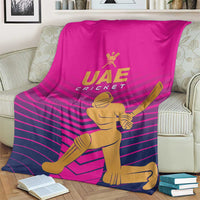 Pink UAE Cricket blanket with gold UAE Cricket text and batsman graphic, shown on couch