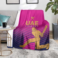Pink UAE Cricket blanket with gold batsman graphic and purple chevron stripes, laid on chair/bench