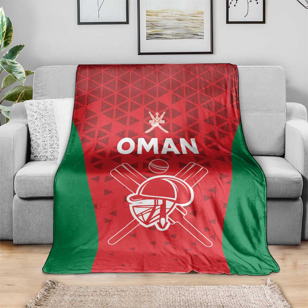 Red and green cricket blanket on a gray sofa with white “OMAN” and helmet bat graphic