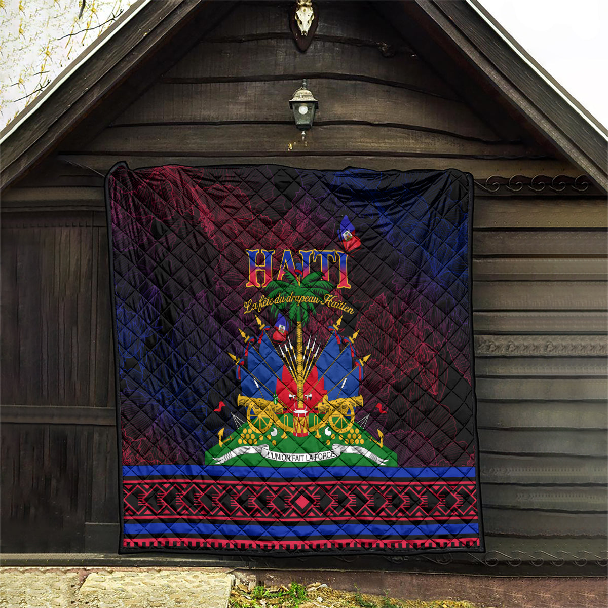 Haitian Flag Day quilt hanging on wall with central Haiti emblem and red/blue patterned border, dark base.
