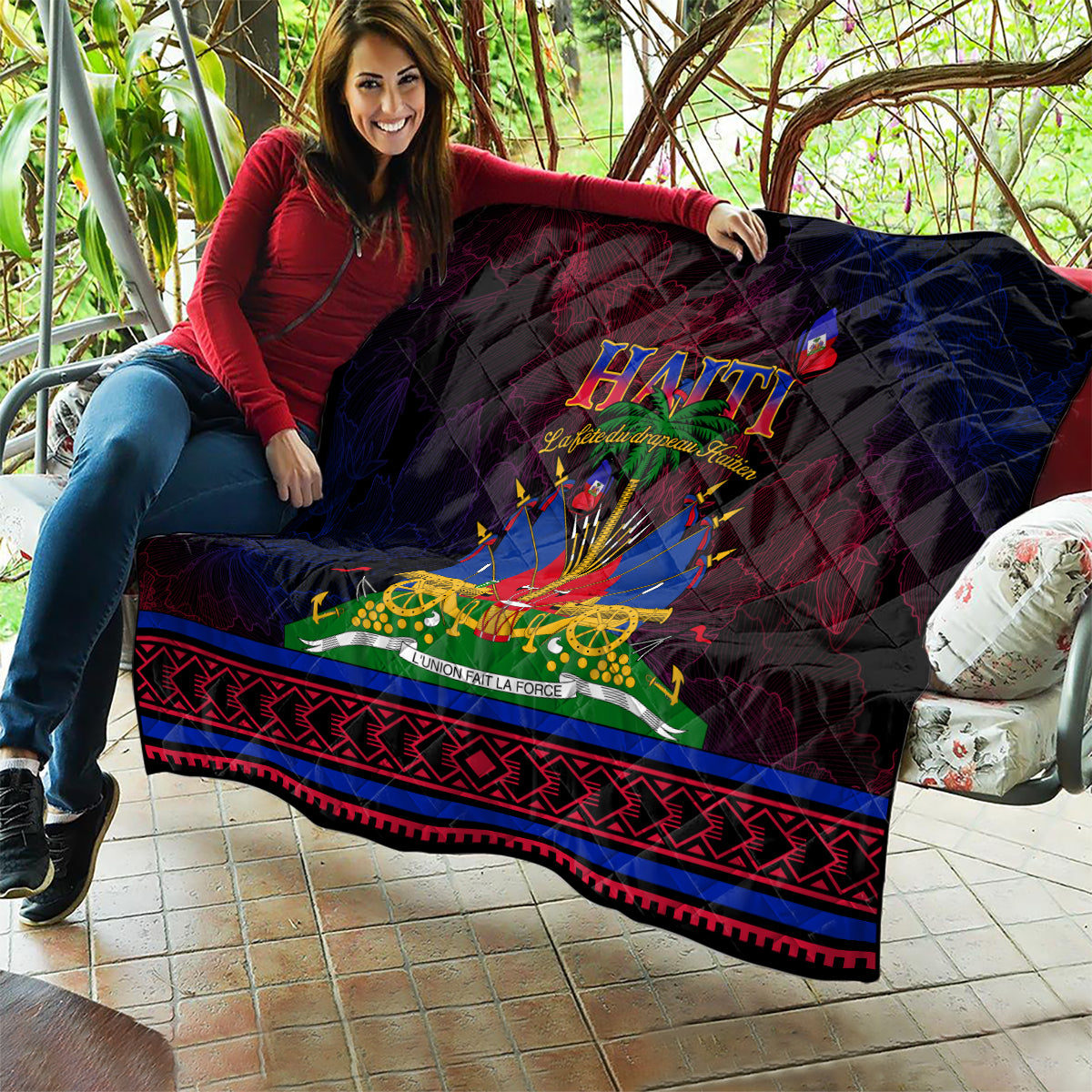Haitian Flag Day quilt held outdoors, showing central Haiti emblem and red/blue patterned hem on dark background.