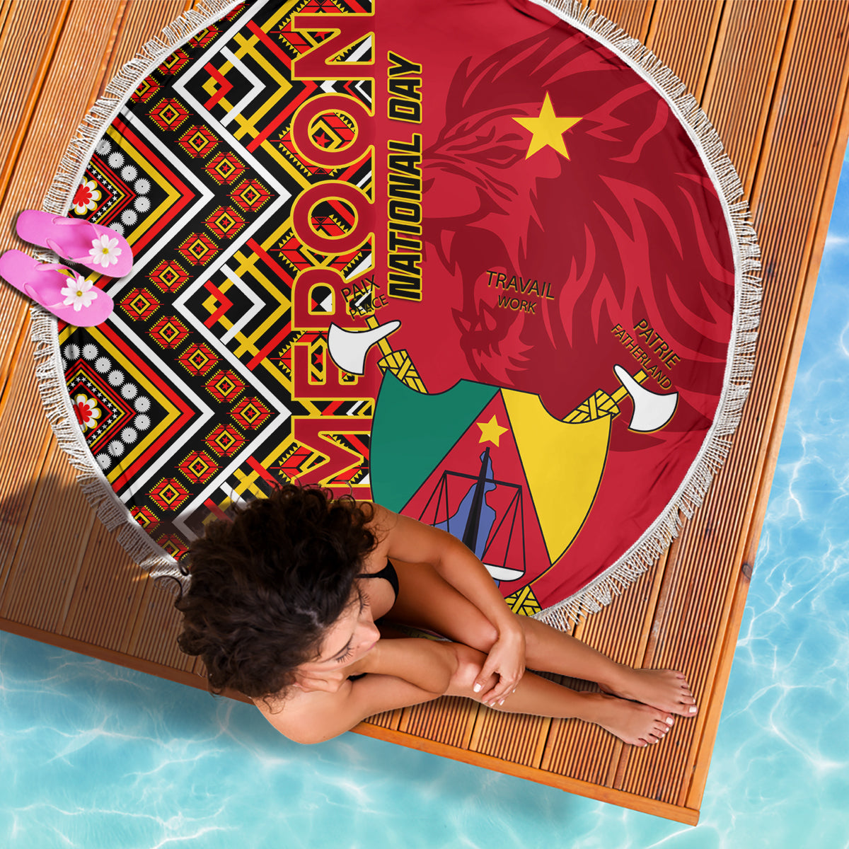 Round fringed beach blanket laid flat, featuring Cameroon coat-of-arms and red Ankara pattern