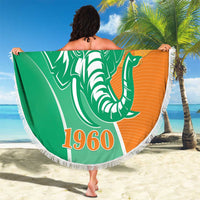 Round beach blanket held outdoors on sand, green/orange elephant graphic with white fringe