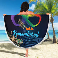 Round navy beach blanket with colorful dragonfly design and white fringe, held up on sand