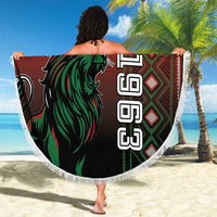 Round red Kenya beach blanket with green lion graphic and white fringe, held on beach