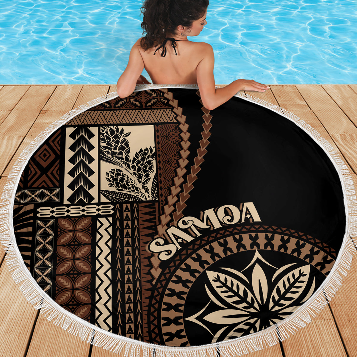 Black round beach blanket with white tassel trim, floral leaf emblem and tapa patterns