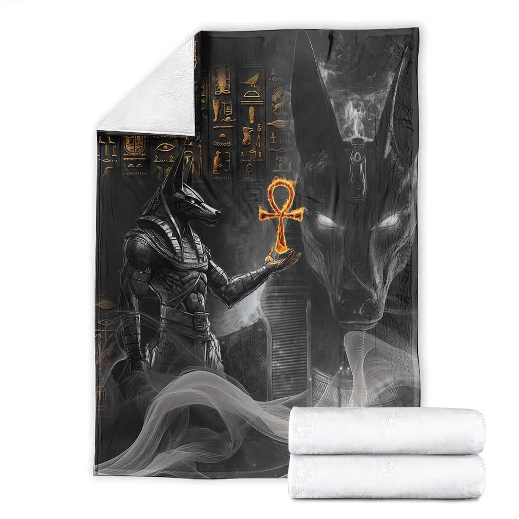 Black Anubis-themed blanket with stylized figures and orange symbol, packaged with rolled white towels