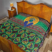Green kanga-style quilt laid on bed with multicolor motifs, central blue figure, and quilted texture
