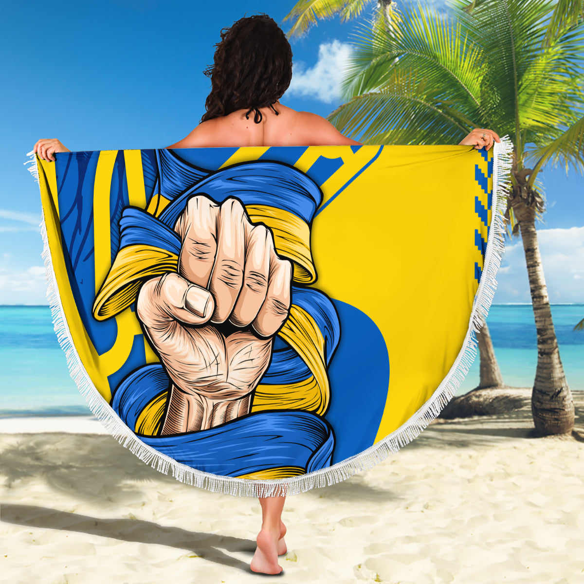 Round beach blanket with blue and yellow trident-fist graphic, fringed edge stretched on beach