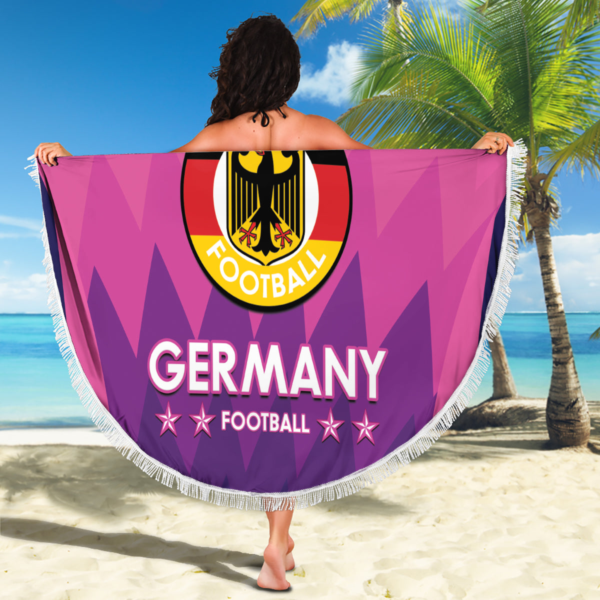Pink round Germany football beach blanket with white fringe, held up on sandy beach