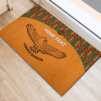 Orange rubber doormat with kente-pattern border, central eagle graphic, and “YOUR TEXT”