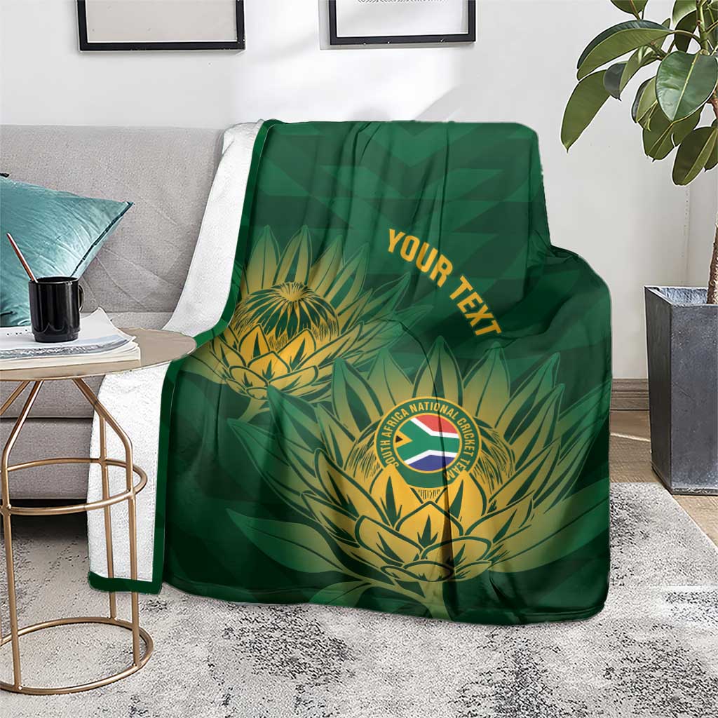 Green cricket blanket with gold crest and tropical leaf artwork, spread over a gray sofa