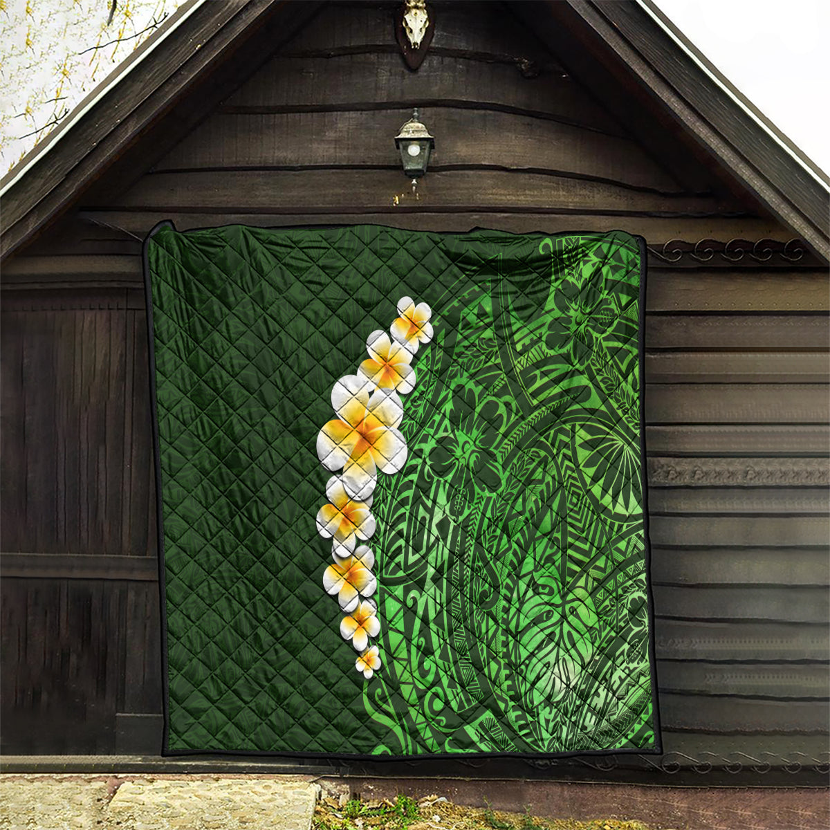 Green quilted blanket hanging indoors, with green Polynesian design and white-yellow plumeria motif.