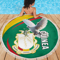 Round fringe beach blanket with Guinea coat of arms and white pigeon graphic on wooden deck.