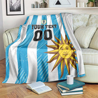Blue and white striped football blanket on couch with Uruguay crest, sun graphic, and 'YOUR TEXT 00'.