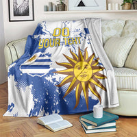 Blue and white Uruguay-themed throw blanket draped on a couch with sun face and “00 YOUR TEXT”