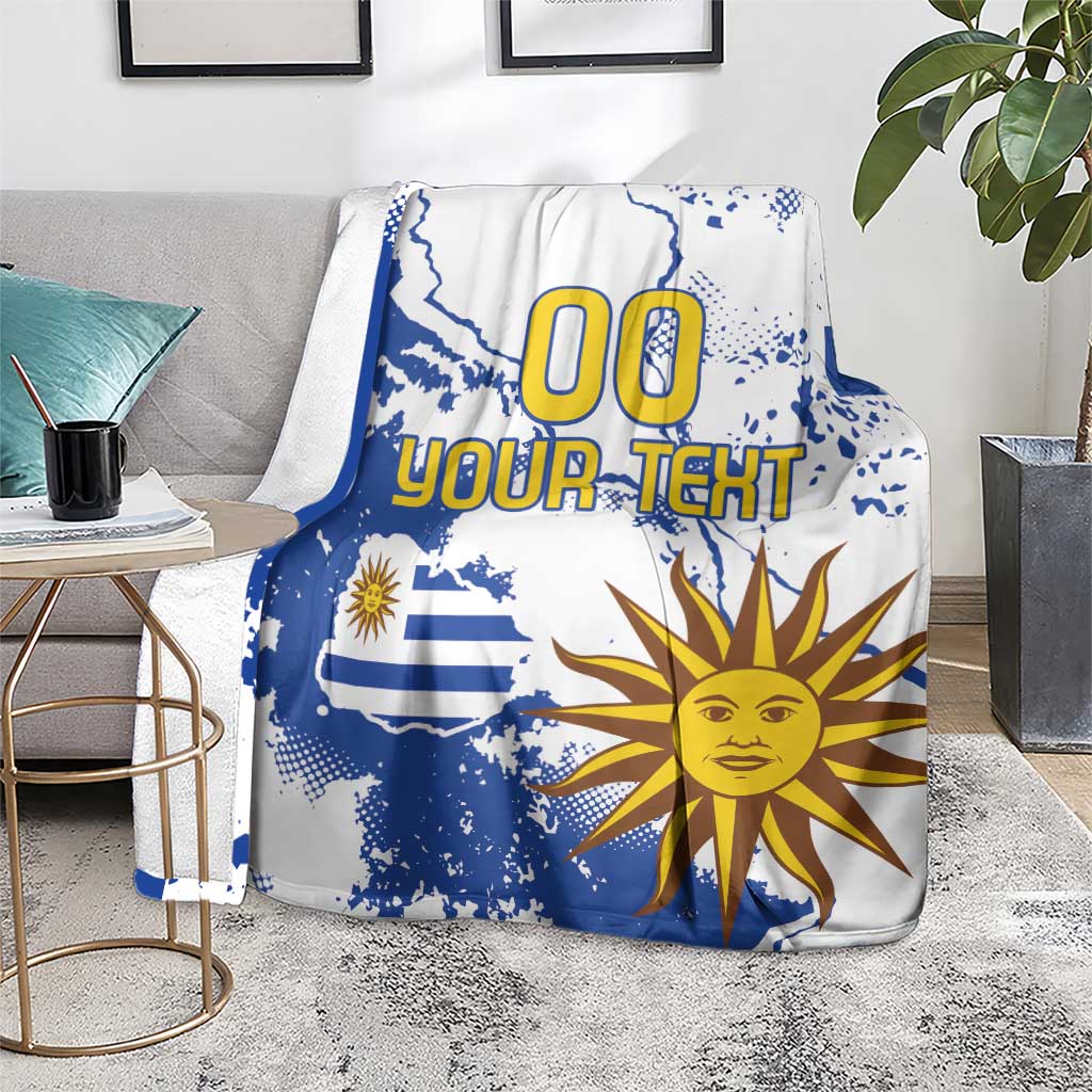 Blue and white Uruguay-themed blanket spread on a gray couch with sun face and “00 YOUR TEXT”