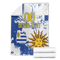 Folded blue and white Uruguay-themed blanket with sun face and “00 YOUR TEXT” lettering