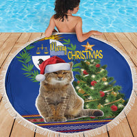 Blue round Christmas beach blanket with white fringe, seated cat in Santa hat by tree