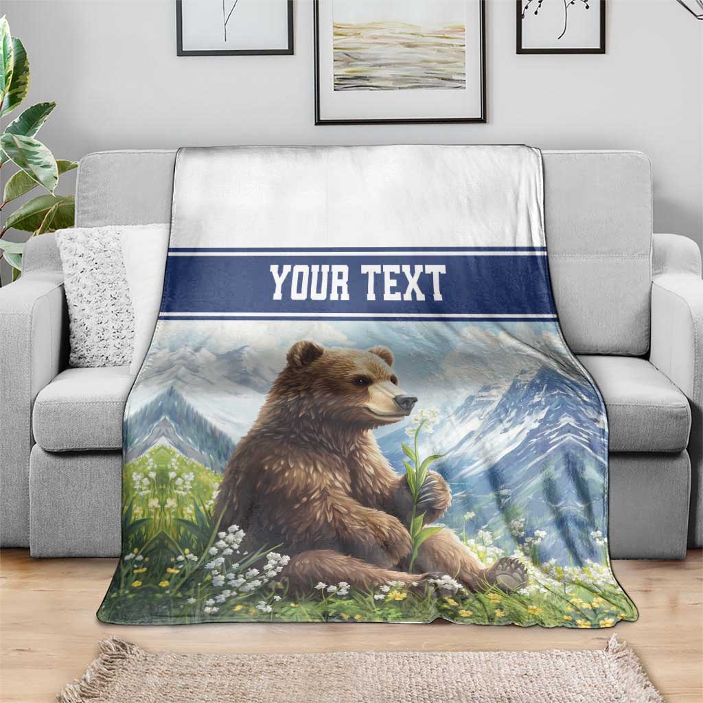 Personalized throw blanket draped over sofa: brown bear, blue mountains, flowers, 'YOUR TEXT'