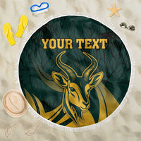Green round beach blanket with fringe edge, gold springbok emblem on leafy background, custom text