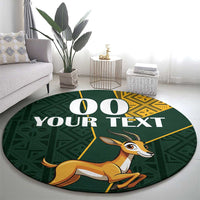 Round area rug in dark green featuring springbok face, “YOUR TEXT” and yellow kente-style trim