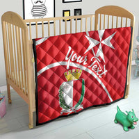 Red quilt blanket with Malta cross, crest shield, and white script on a baby bed