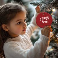 Red round ornament with 'JESUS WON' text, hanging on a Christmas tree branch.