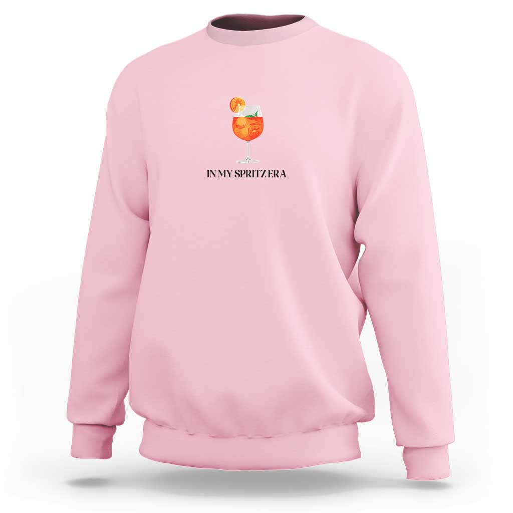 Light pink crewneck sweatshirt with small orange drink graphic and “IN MY SPRITZ ERA” text