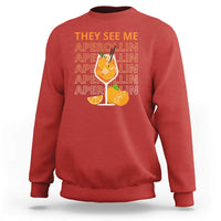 Red sweatshirt with “THEY SEE ME” Aperol-lin text and citrus cocktail graphic on back