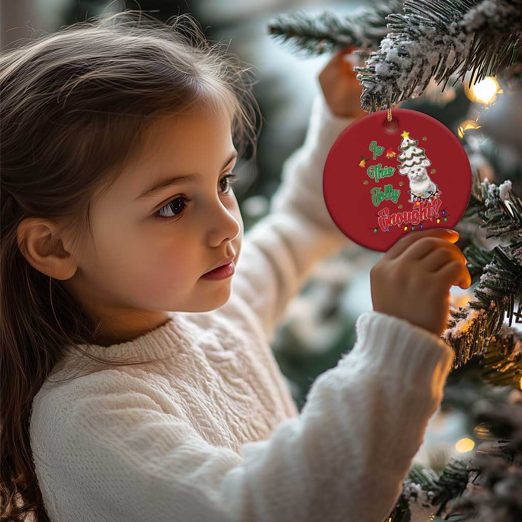 Red round cat Christmas ornament held by a child near an evergreen tree.
