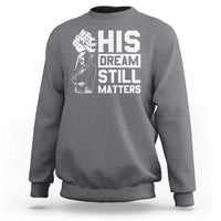 Charcoal gray crewneck sweatshirt with white "HIS DREAM STILL MATTERS" graphic front.