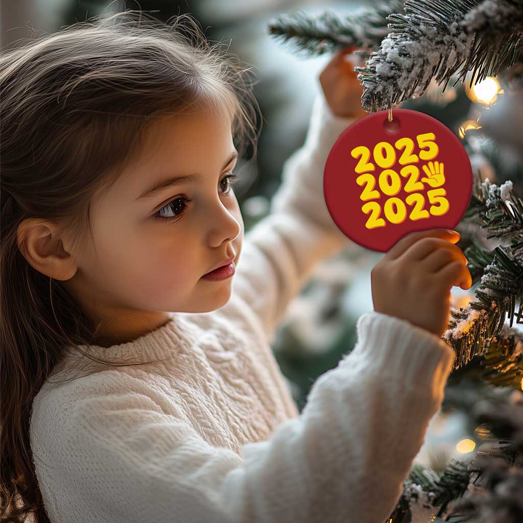 Red round 2025 ornament with yellow “2025” and handprint icon, hanging on a tree