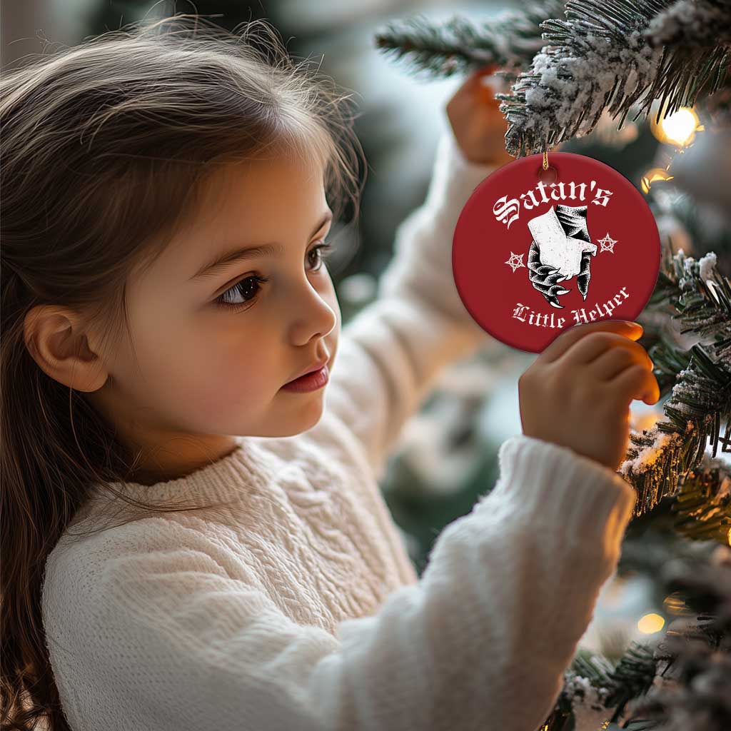 Red round Satan's Little Helper ornament held by a child near the tree