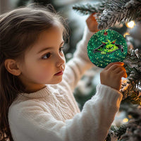 Green circular cat Christmas ornament held by a child near a decorated Christmas tree.