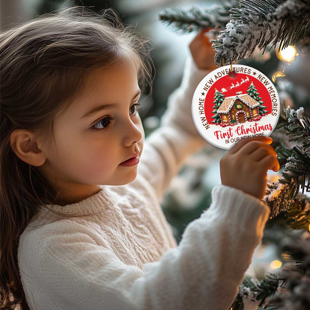 Red round Christmas ornament held near a Christmas tree, featuring a decorated house and “First Christmas 2024”.