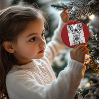 Red round Christmas ornament with selfie-style reindeer graphic, held near a decorated tree
