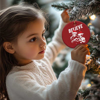Red round “BELIEVE” Christmas ornament with alien and Bigfoot, held near a tree